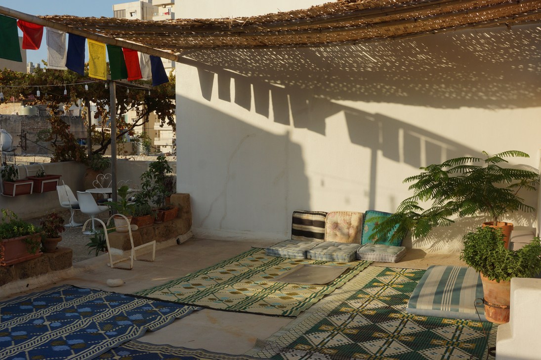Bohemian rooftop terrace with colorful prayer flags, patterned rugs, cushioned seating, potted plants and white pergola at Beit el Nessim.