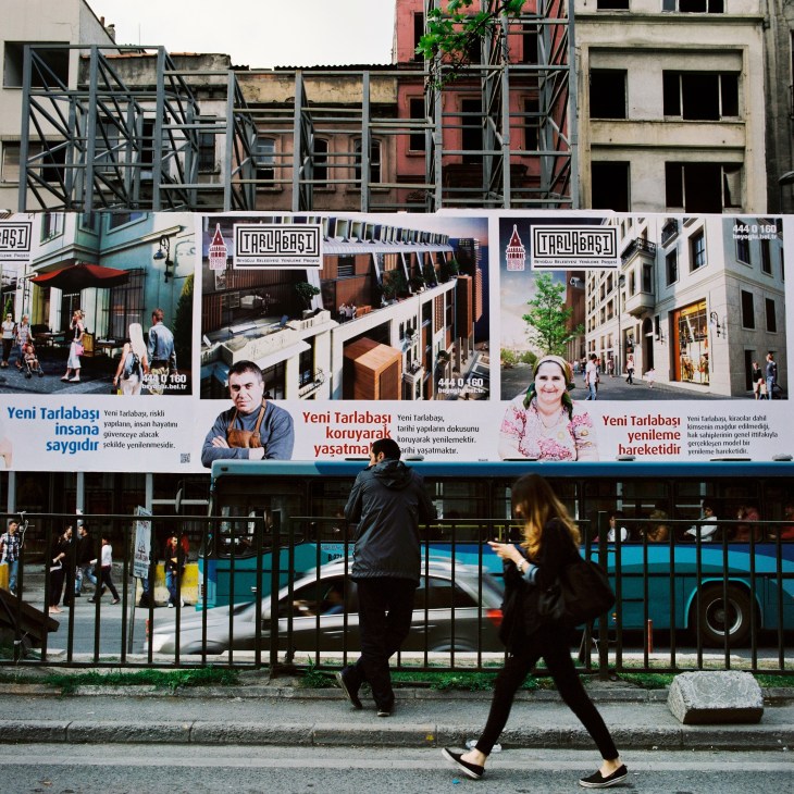 Posters announcing new developments in the area surrounding Taksim Square, with derelict buildings behind and pedestrians passing by.