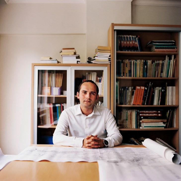 Tayfun Kahraman, chairman of the Turkish Chamber of Urban Planners, sits at a desk with architectural plans in his office surrounded by bookshelves.