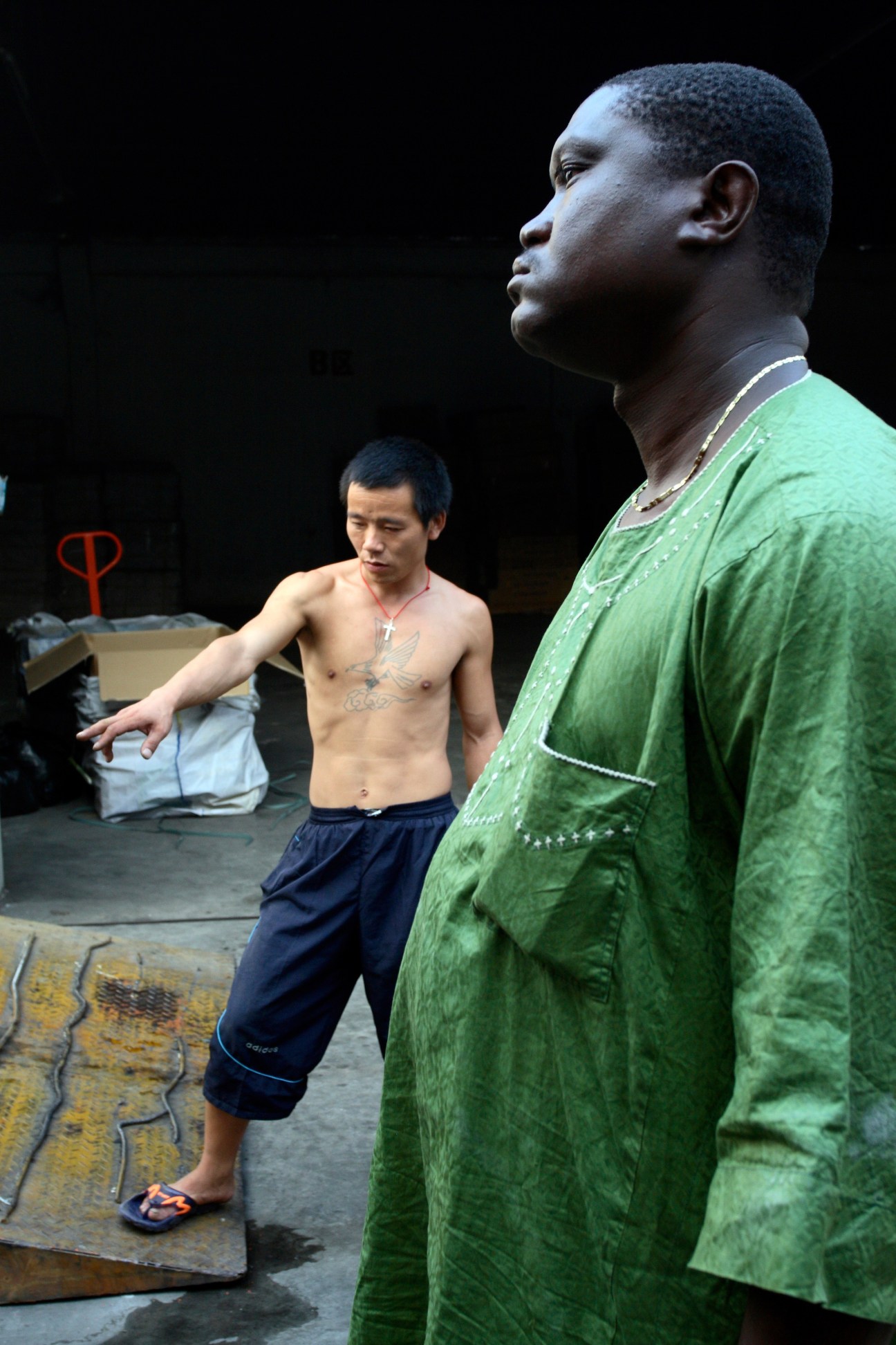 An African man in a green shirt stands in a warehouse while a shirtless Chinese worker handles cargo nearby.