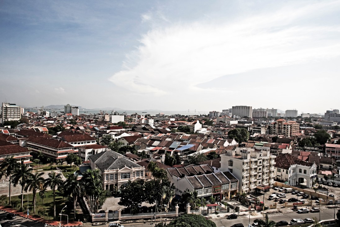 Aerial view of George Town showing colonial shophouses, palm trees, and modern buildings with a bridge visible across the water.