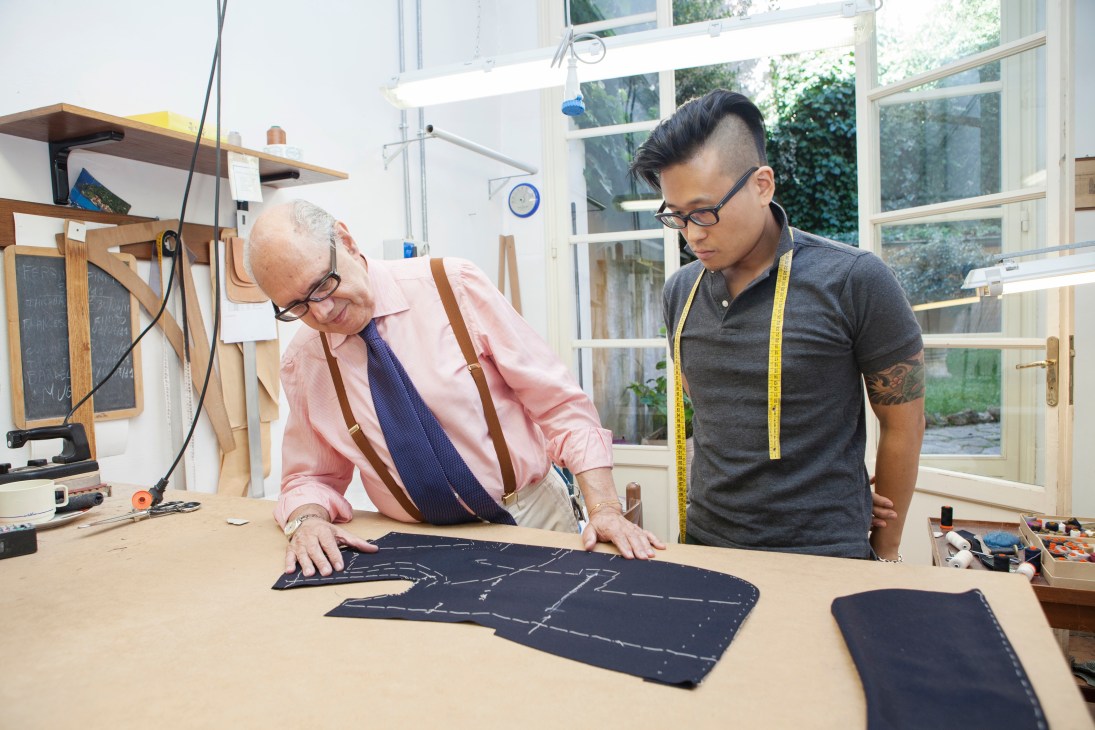 Antonio Liverano inspects apprentice Hojun Choi's tailoring work on a navy fabric pattern at a wooden workbench.