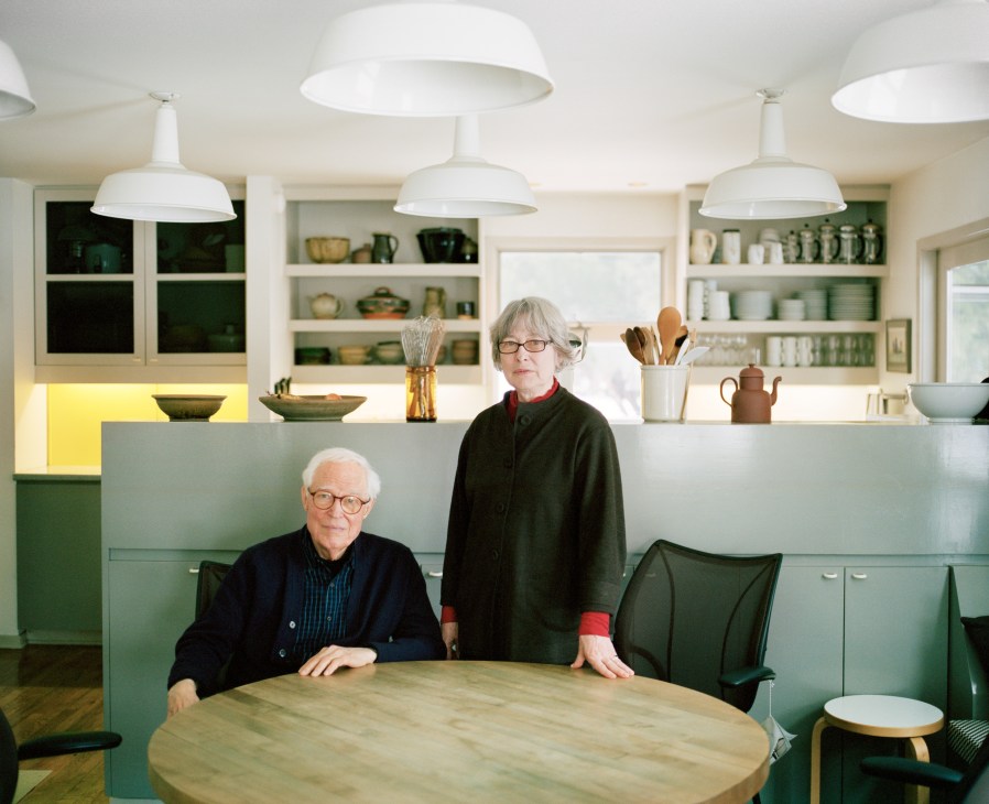 Industrial designer Niels Diffrient seated and his wife Helena Hernmarck standing beside a round table in their kitchen.