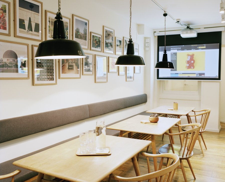 Monocle Café downstairs seating area with wooden tables, Ercol chairs, gray bench seating, framed artwork, pendant lights and projection screen.