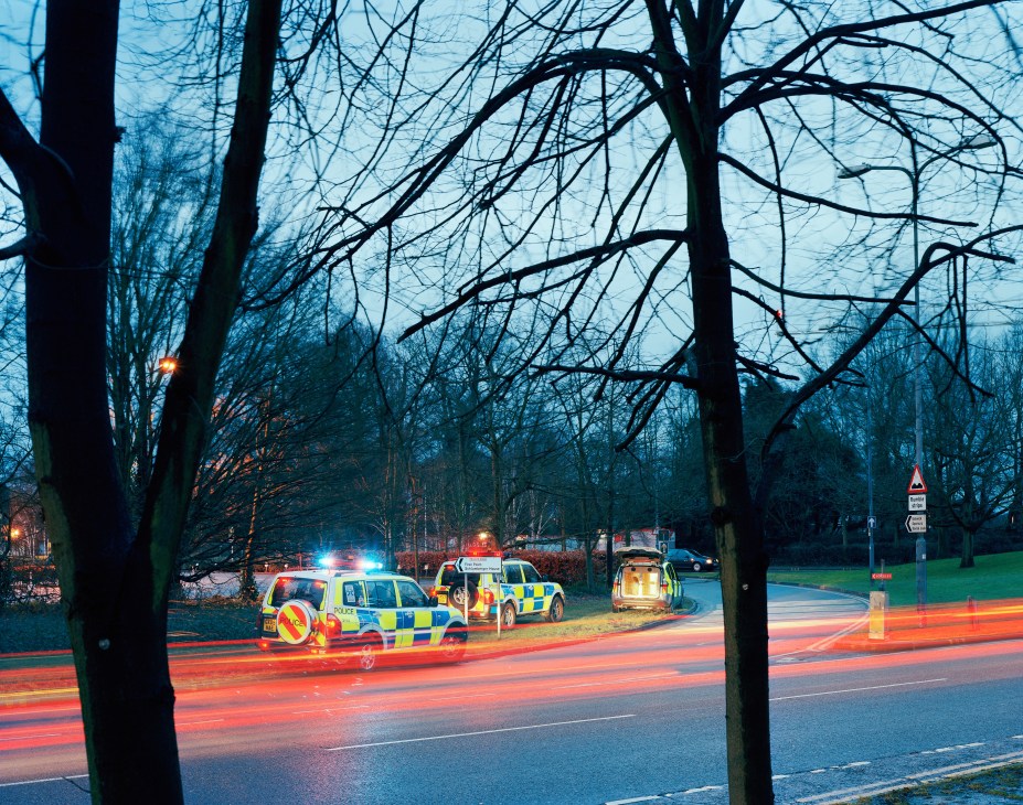 Police vehicles with flashing lights at a dawn checkpoint near bare trees and a road.