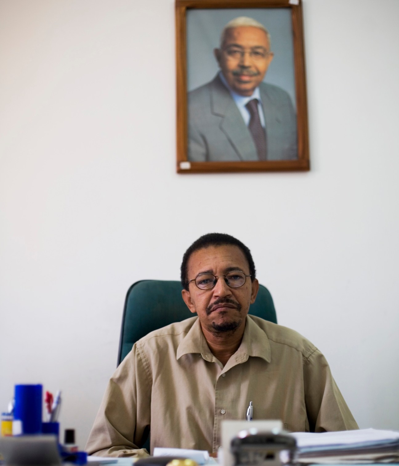 Alfredo Santos, director of InforPress agency, seated at desk with framed photograph of President Pedro Pires on wall behind.