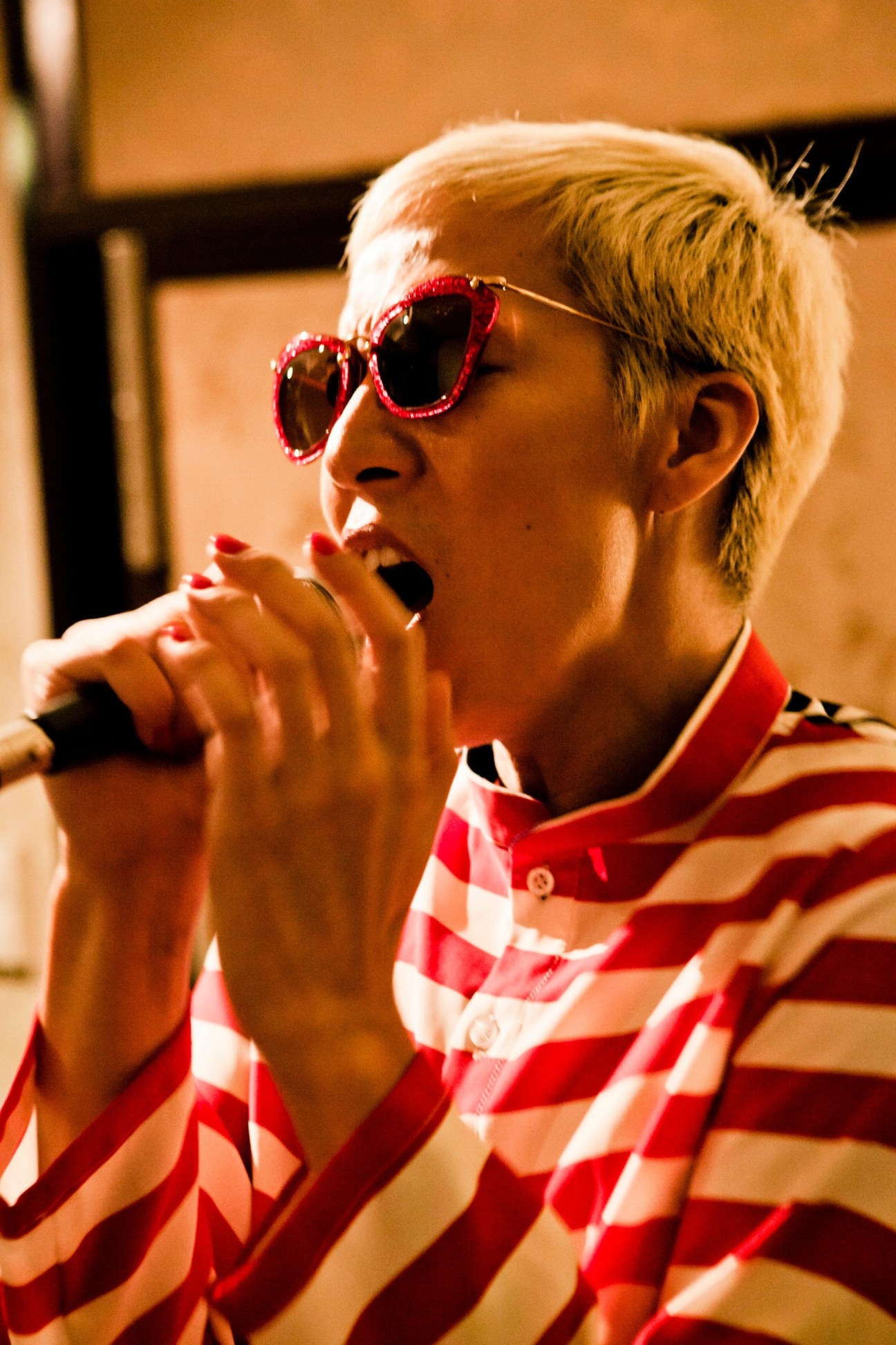 Gene Kasidit wearing red sequined heart-shaped sunglasses and a red and white striped shirt, singing into a microphone.