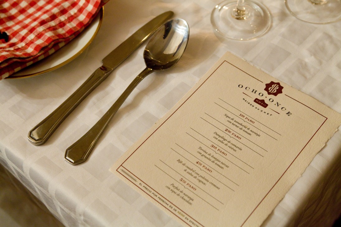 Ocho Once menu card on white tablecloth with cutlery and red checkered napkin.