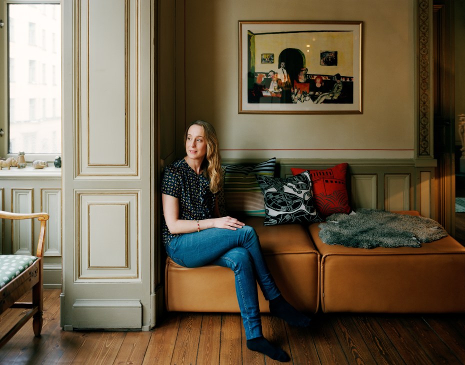 Karin Collste sits on a tan leather sofa in her apartment's living room with painted ceiling and framed artwork.
