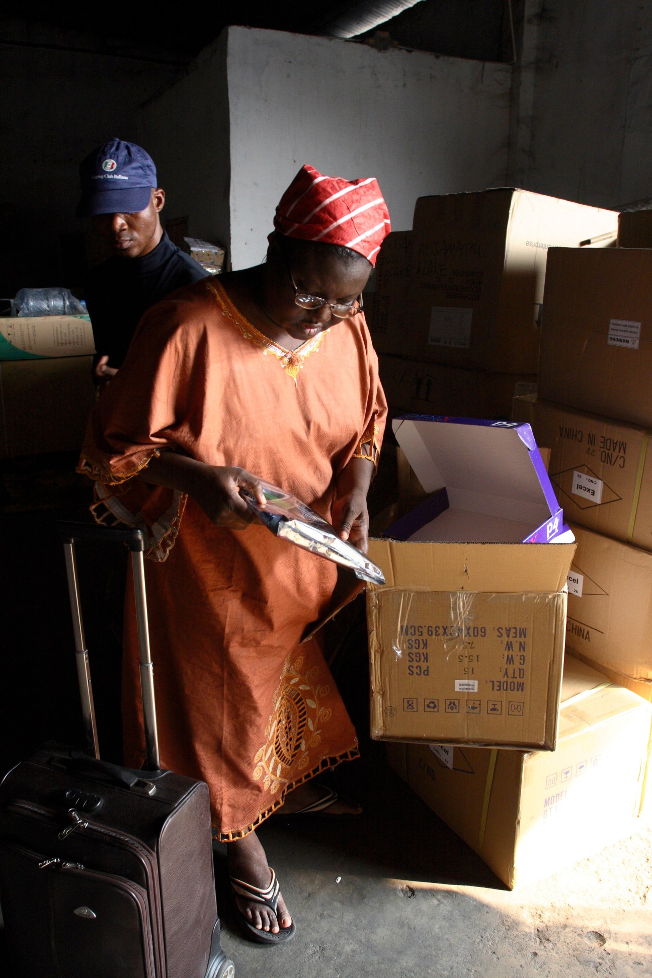 Two people inspect cardboard boxes of goods at a warehouse, with one woman opening a box in an orange dress and red headwrap.