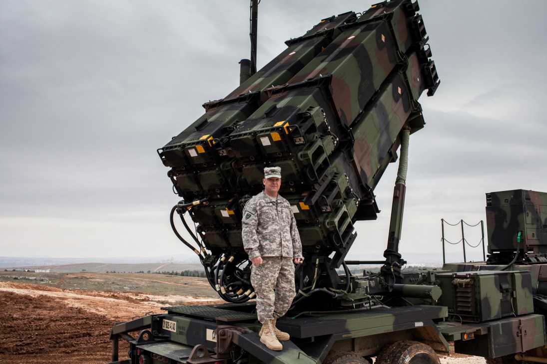 Colonel John Wanat in military uniform stands in front of a PAC-3 Patriot missile launcher on a hillside in Turkey.