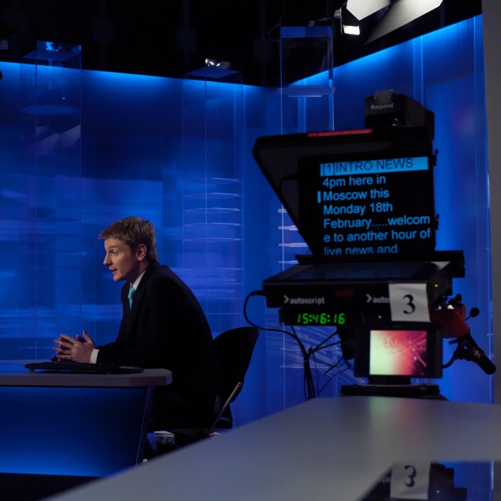 Sports correspondent Peter Oliver sits at a news desk in a blue-lit Russia Today studio during a broadcast.