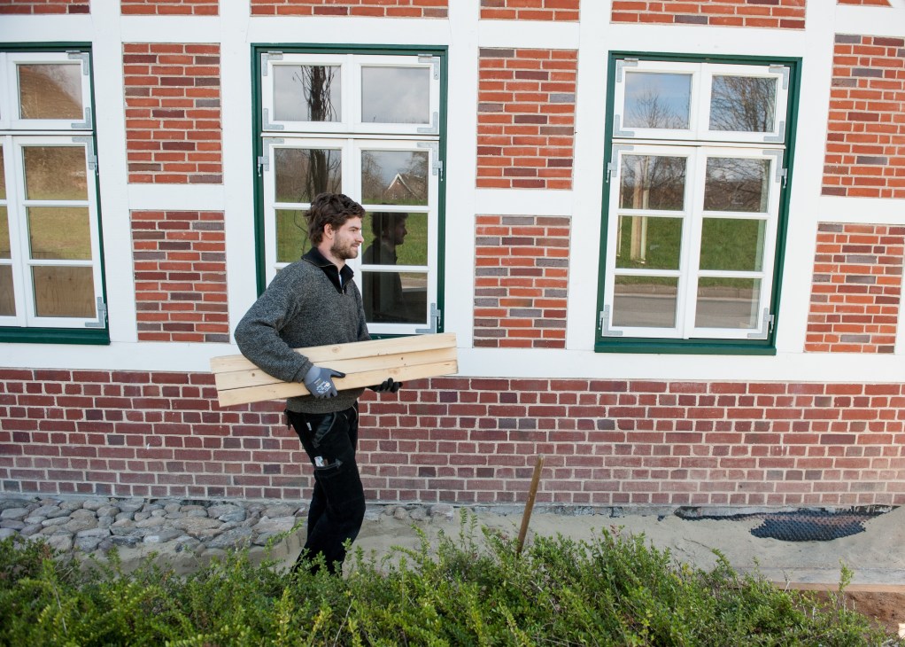 Apprentice carpenter Jonas Klann carries a wooden plank in front of a brick building with green-trimmed windows during a restoration project.