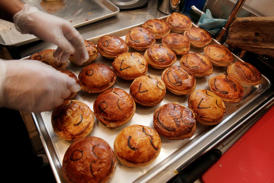 A baker's gloved hand reaches for a freshly baked Australian meat pie on a metal tray lined with parchment paper.