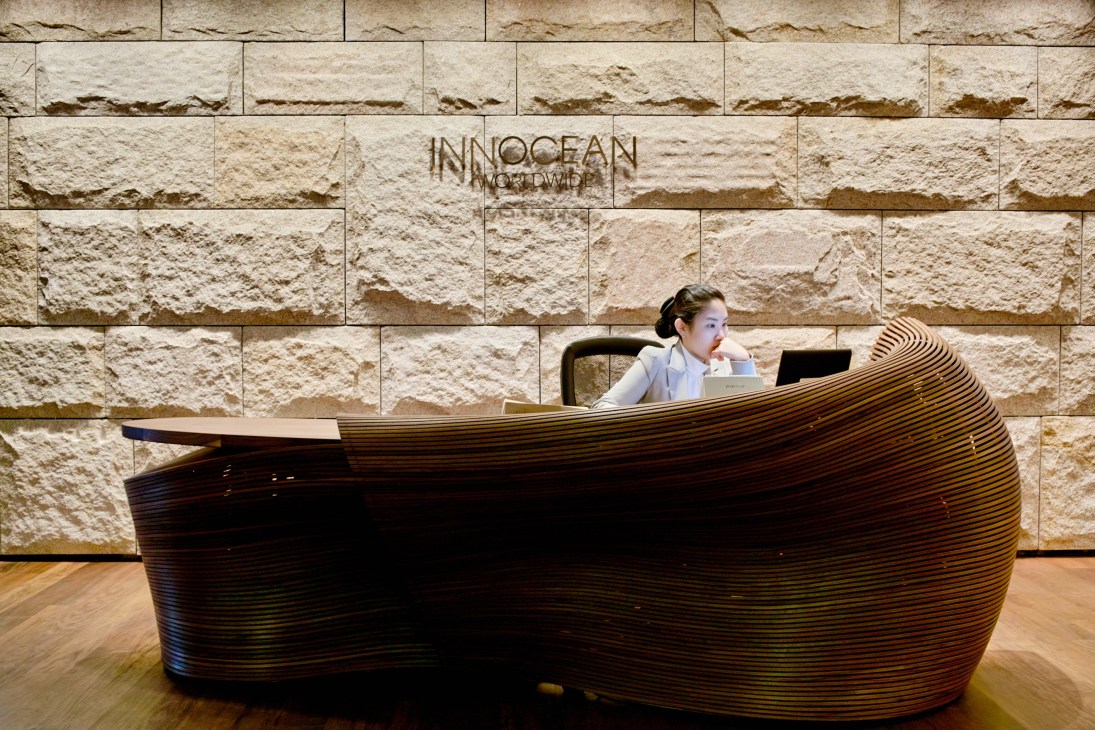 Receptionist at Bae Se Hwa's sculptural walnut desk at Innocean's headquarters with stone wall behind.
