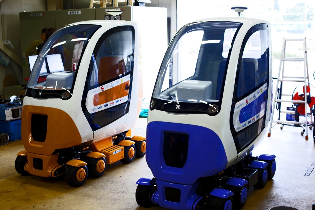 Two compact electric vehicle prototypes with white roofs and colored bodies displayed in a laboratory setting.