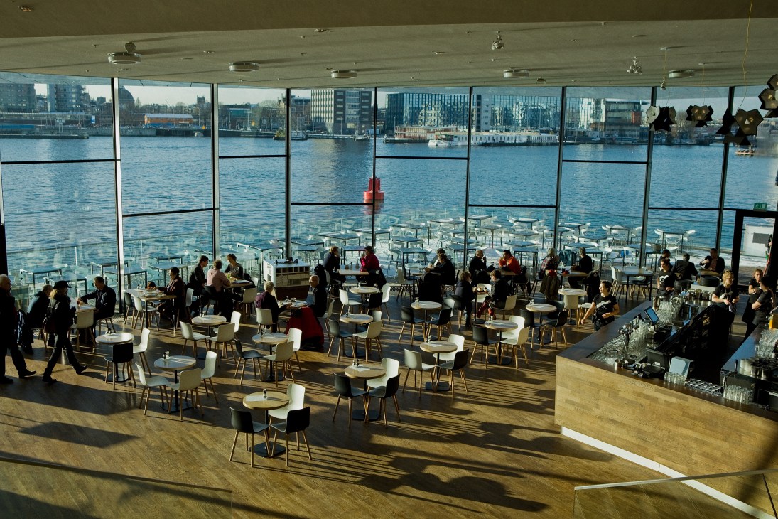 Modern waterside restaurant with floor-to-ceiling windows overlooking Amsterdam's harbor and cityscape.