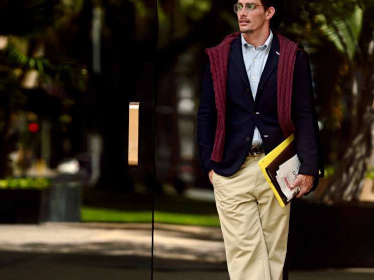 Man wearing navy blazer, cream trousers, burgundy cardigan and glasses holding a yellow-trimmed document case.