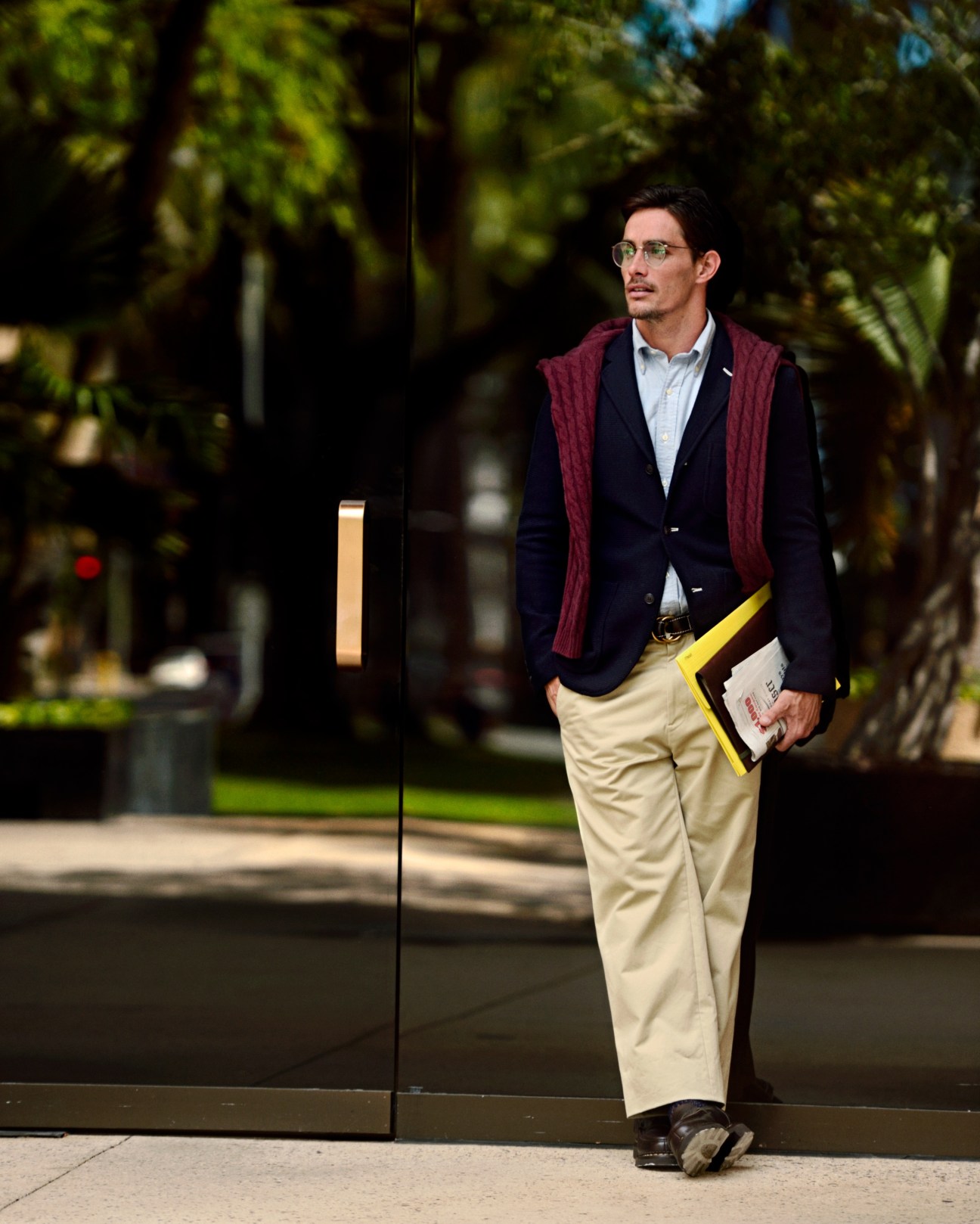 Man wearing navy blazer, cream trousers, burgundy cardigan and glasses holding a yellow-trimmed document case.