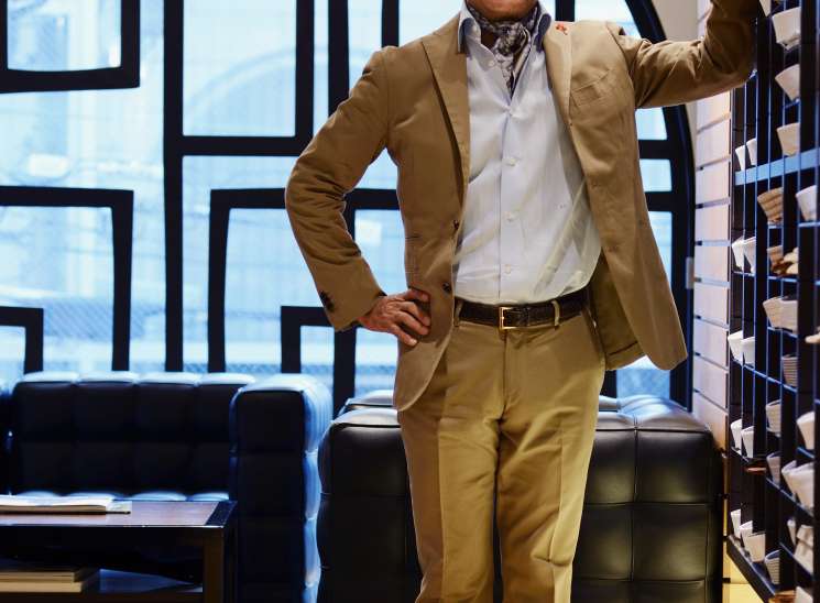 Junji Tajima, owner of Strasburgo, smiling in his Tokyo menswear shop wearing a tan blazer and brown loafers.