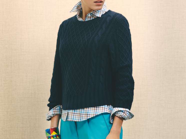 Woman wearing dark quilted jumper, checked shirt, turquoise trousers, colorful bracelets and holding a green bag.
