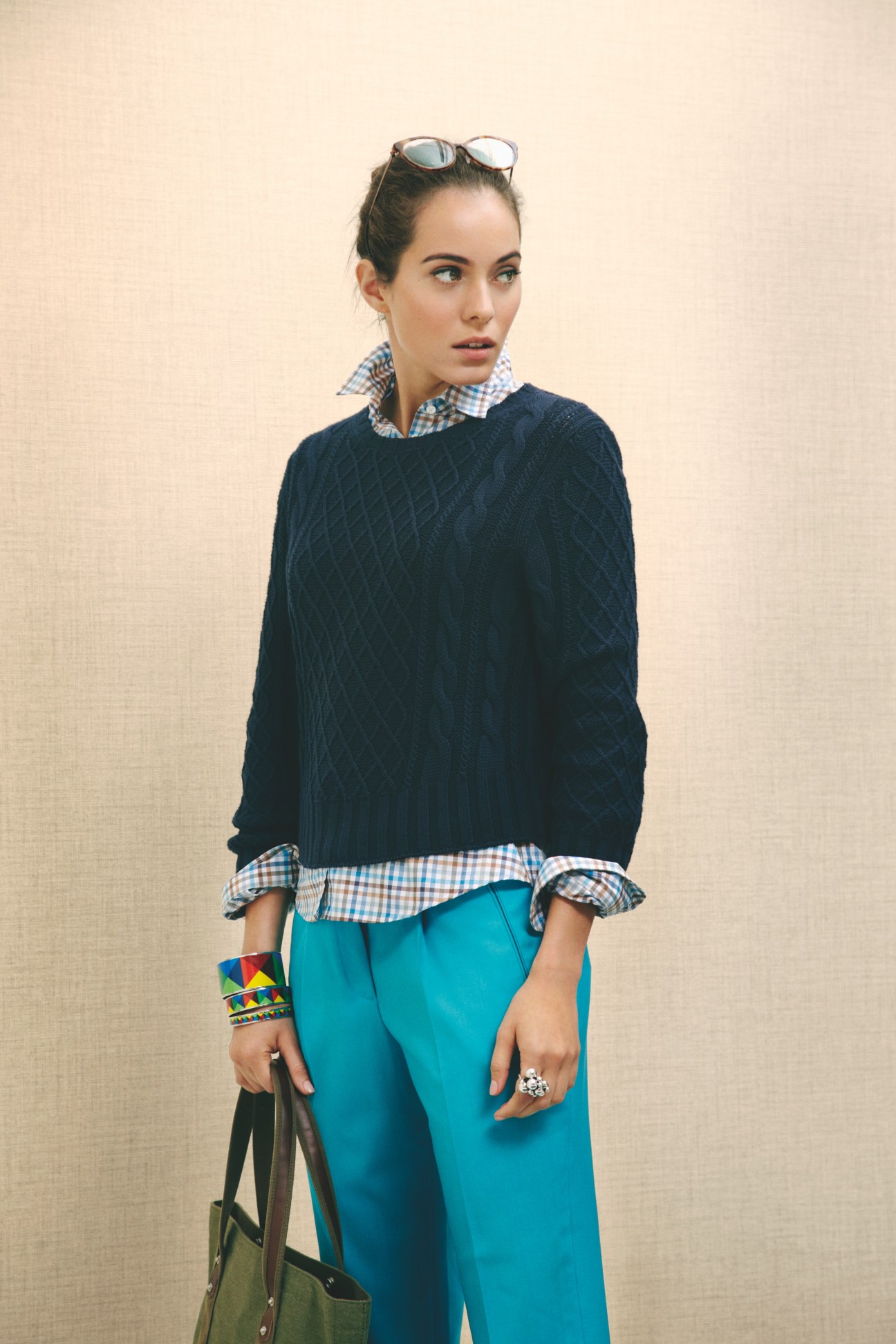 Woman wearing dark quilted jumper, checked shirt, turquoise trousers, colorful bracelets and holding a green bag.