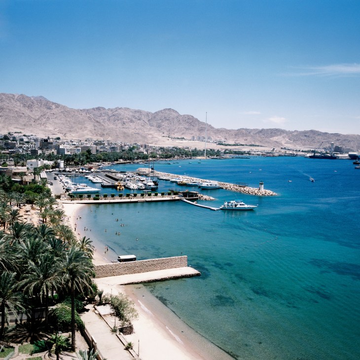 Aerial view of Aqaba's seafront with beach, marina, port facilities, and desert mountains beyond.