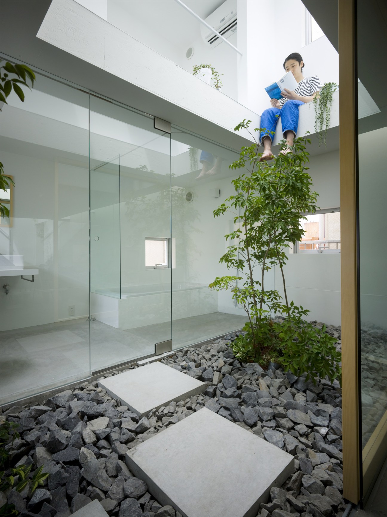 Person reading on an overhang overlooking an indoor courtyard with plants and stone path.