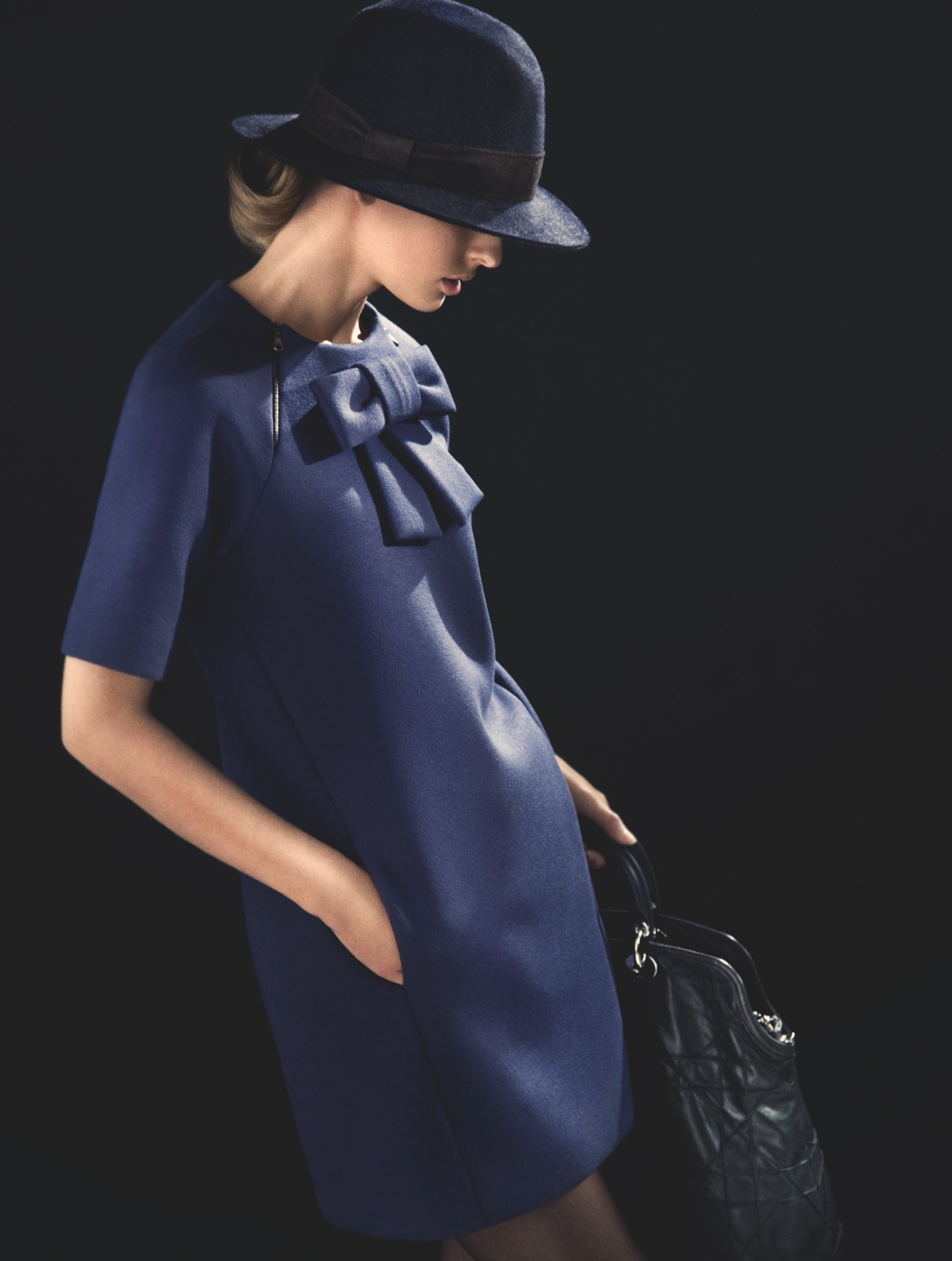 Model wearing navy Mulberry dress with bow detail, Loro Piana hat, and Dior bag against dark background.