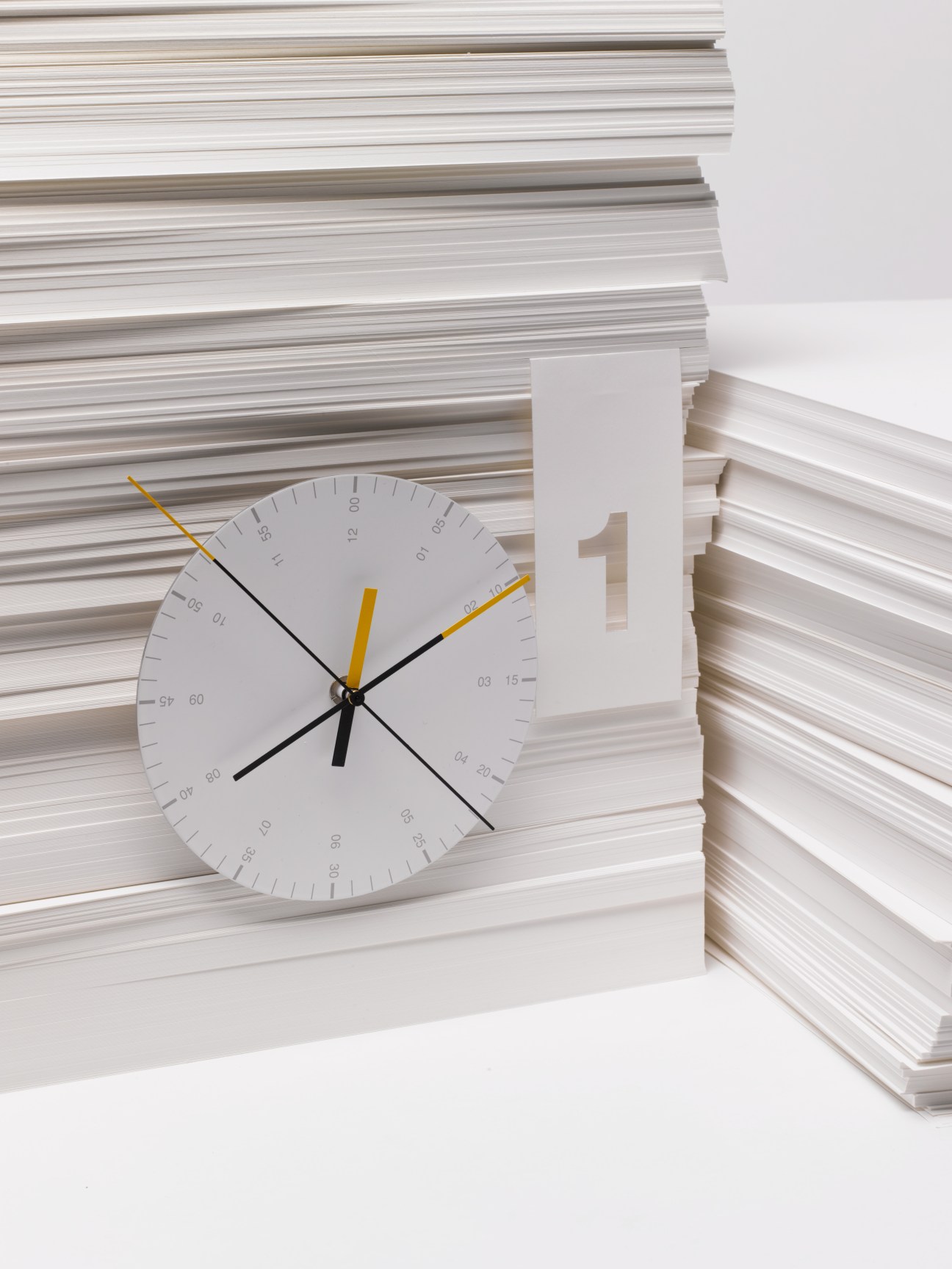 Hundreds Tens Units wall clock with aluminium face and yellow and black hands surrounded by stacked white papers.