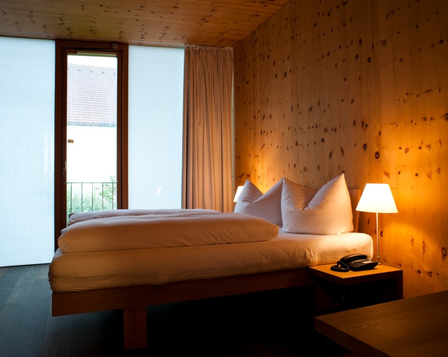 Minimalist bedroom with wooden ceiling and spotted walls, white bedding, and French doors opening to balcony.