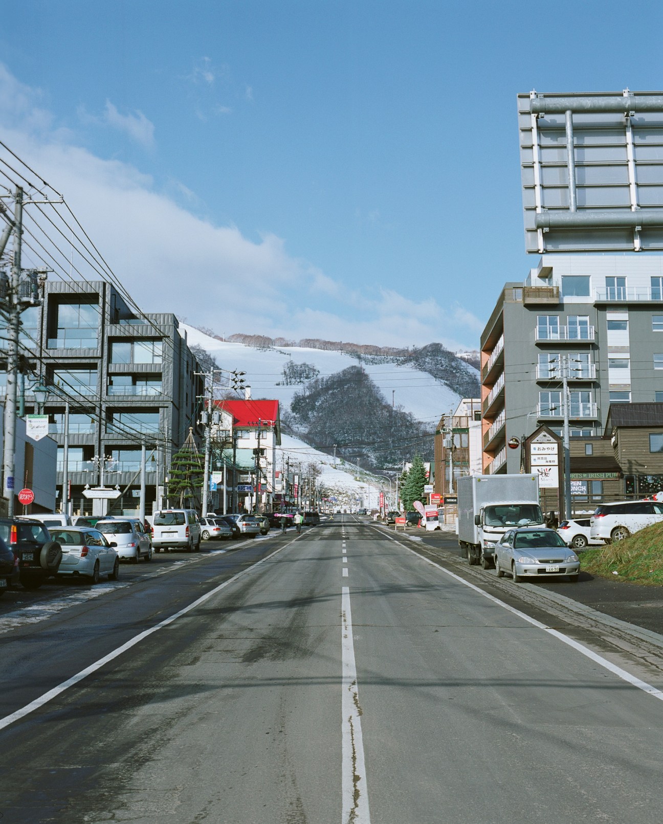Hirafu main street lined with modern apartment buildings and shops, with snow-covered ski slope visible in the distance.