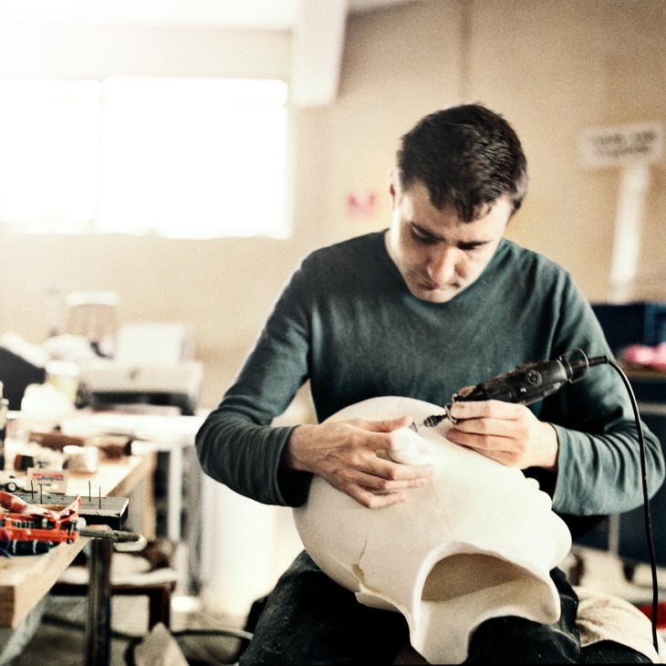 An assistant uses an airbrush tool to detail a white puppet head in a workshop studio.