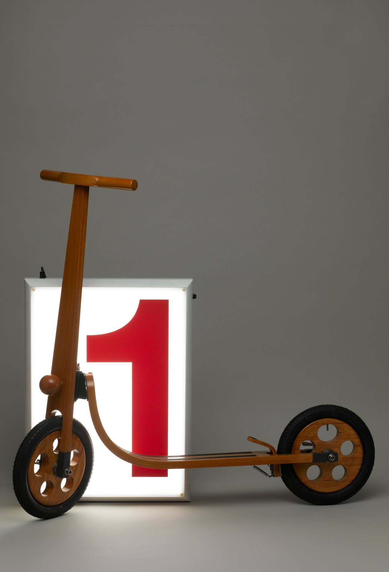Wooden kick-scooter with brown handcrafted wooden frame and black wheels next to illuminated number one.