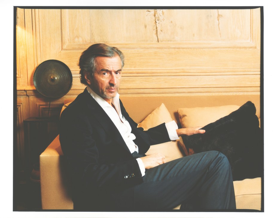 Bernard-Henri Lévy sitting on a beige sofa in a wood-panelled room, holding a book.