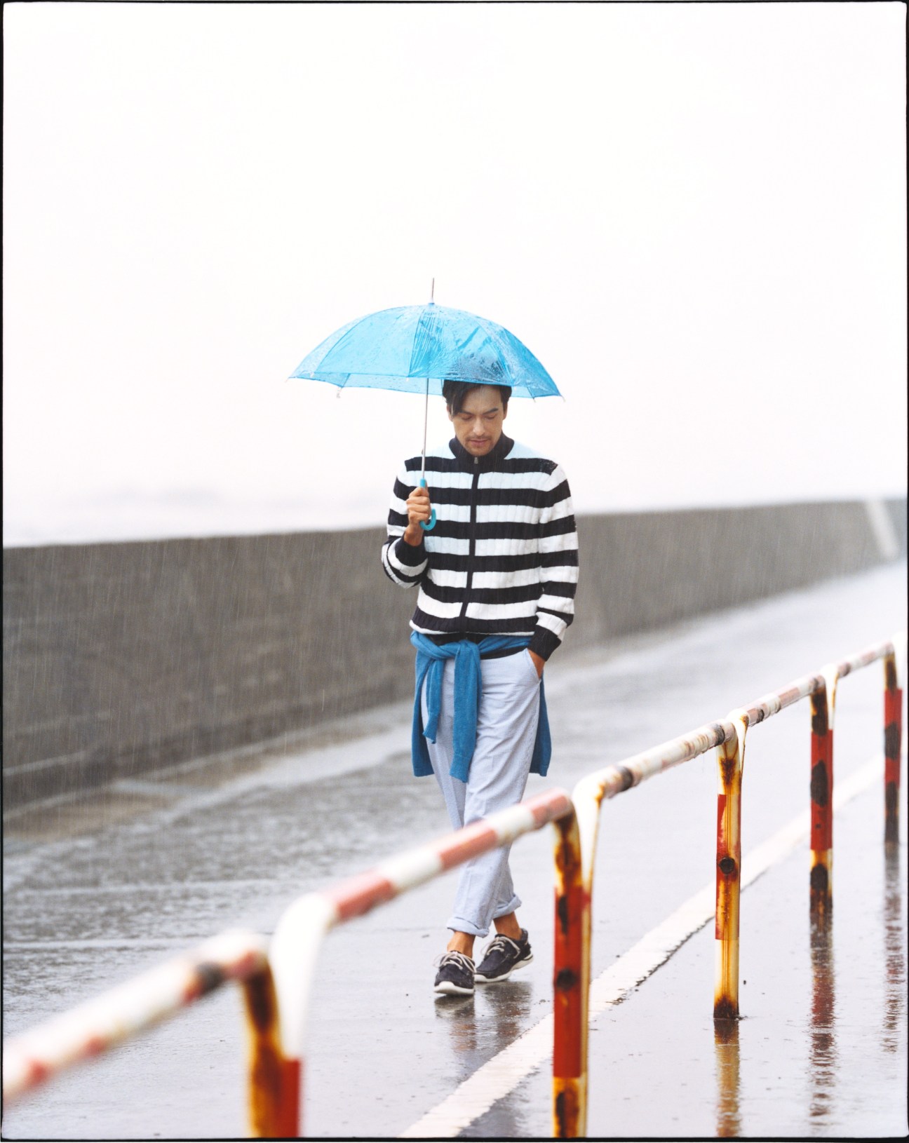 Model wearing Brooks Brothers sweater and trousers with striped jumper, holding blue umbrella on wet boardwalk in rain.