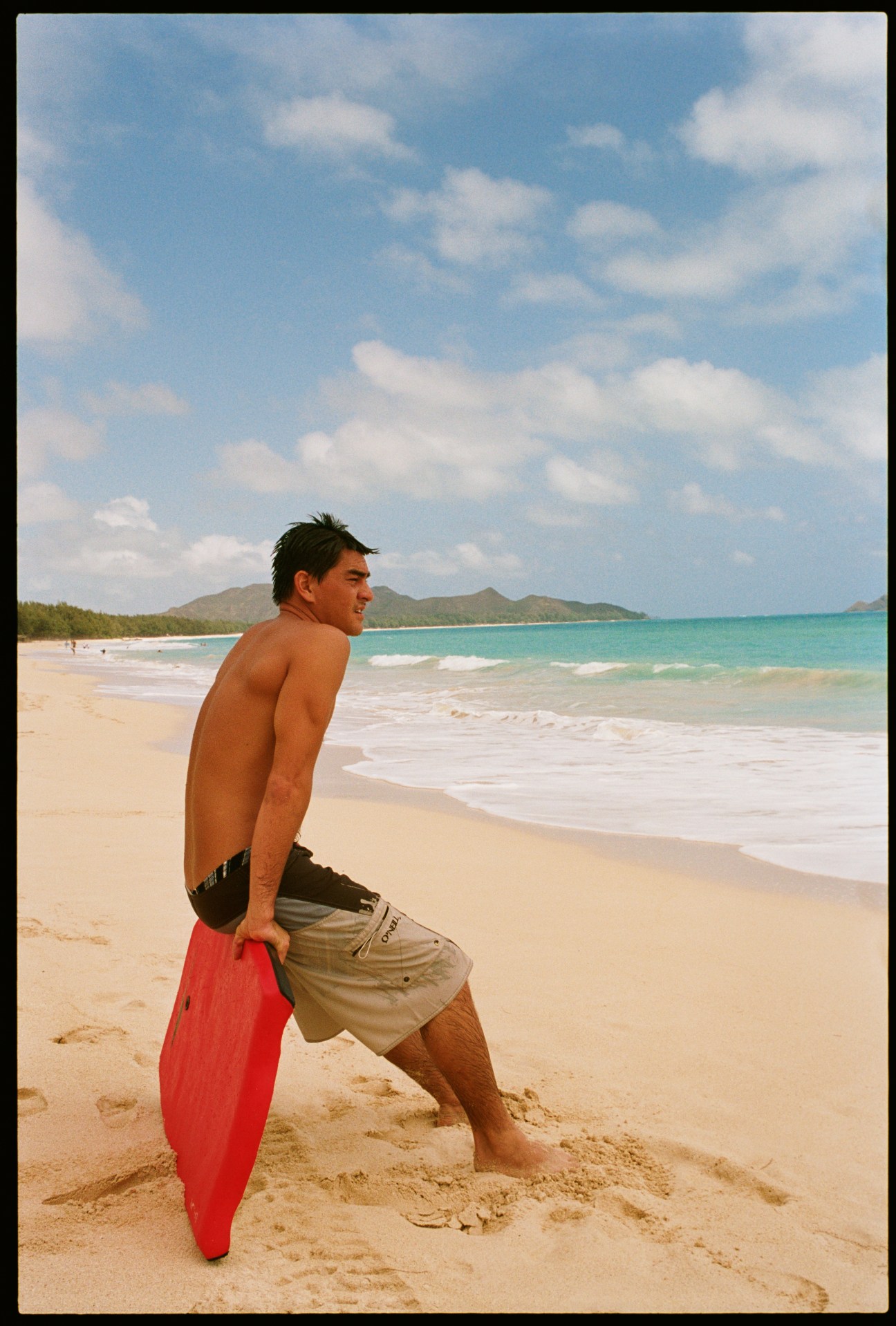A young man holds a red bodyboard while standing on a sandy beach with turquoise waters and mountains in the background.