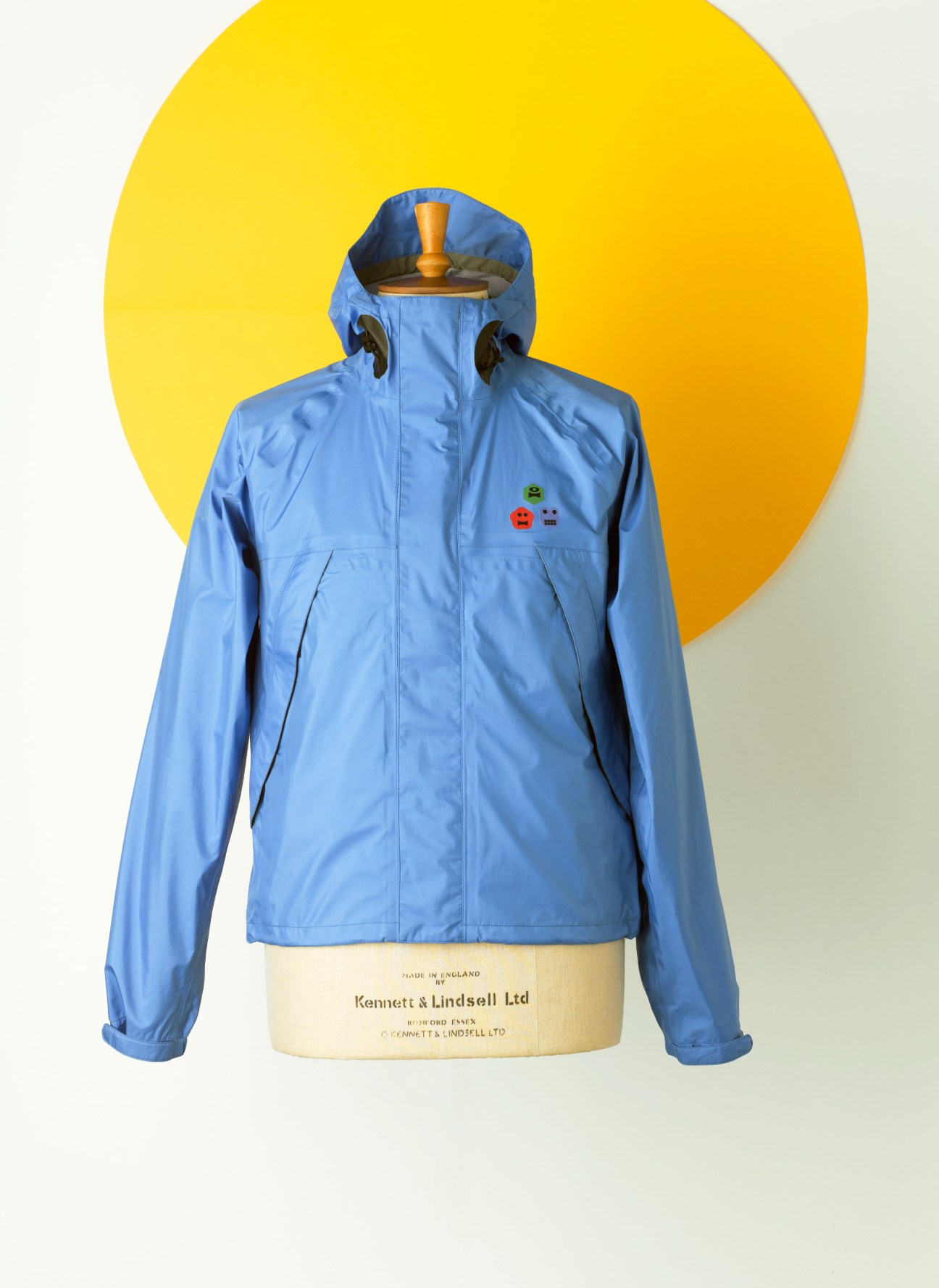 Blue waterproof Aspesi parka with cute face illustration on chest against yellow circular background.