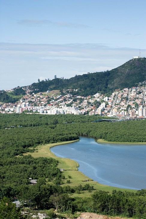Aerial view of Florianópolis with buildings nestled against forested hills, a blue lagoon in the foreground, and a communication tower visible on the hillside.
