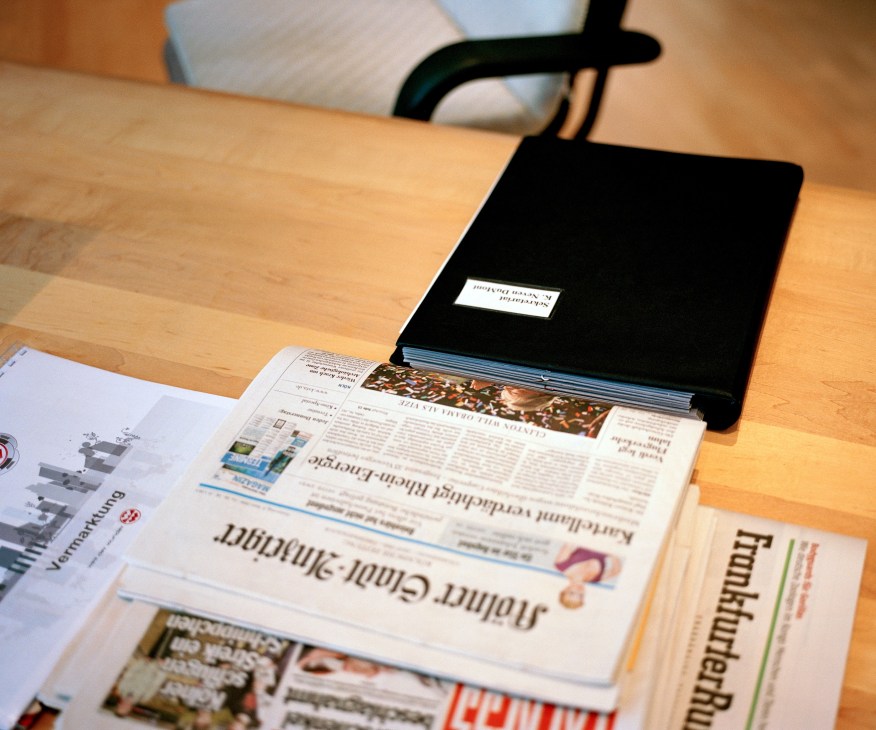 Kölner Stadt-Anzeiger newspaper displayed on a wooden desk with other publications and a black chair visible.