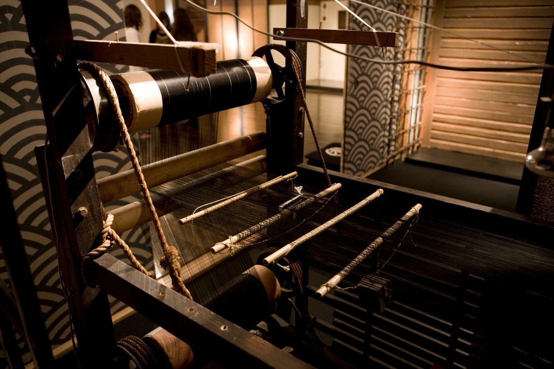 A Meiji-era wooden loom with spindles and thread guides displayed in a traditional Japanese setting.