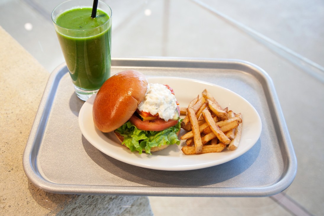 A burger with white sauce, lettuce and tomato on a red bun, served with fries and a green smoothie.