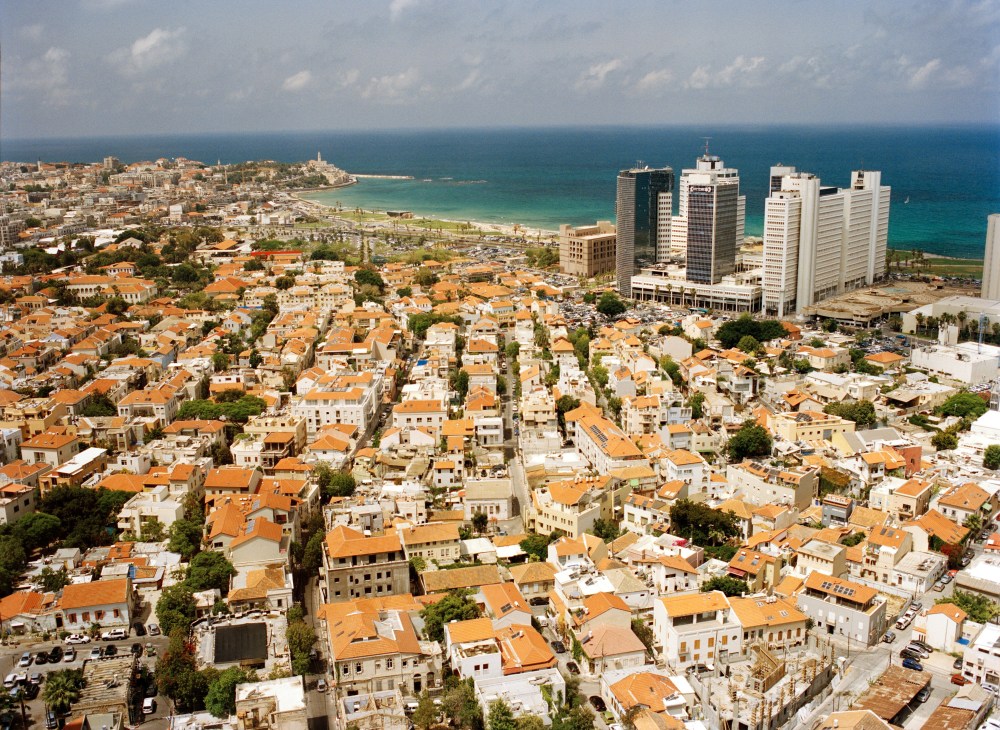 Aerial view of Tel Aviv's Jaffa district showing orange-roofed buildings, the Mediterranean coastline, and modern high-rise towers.