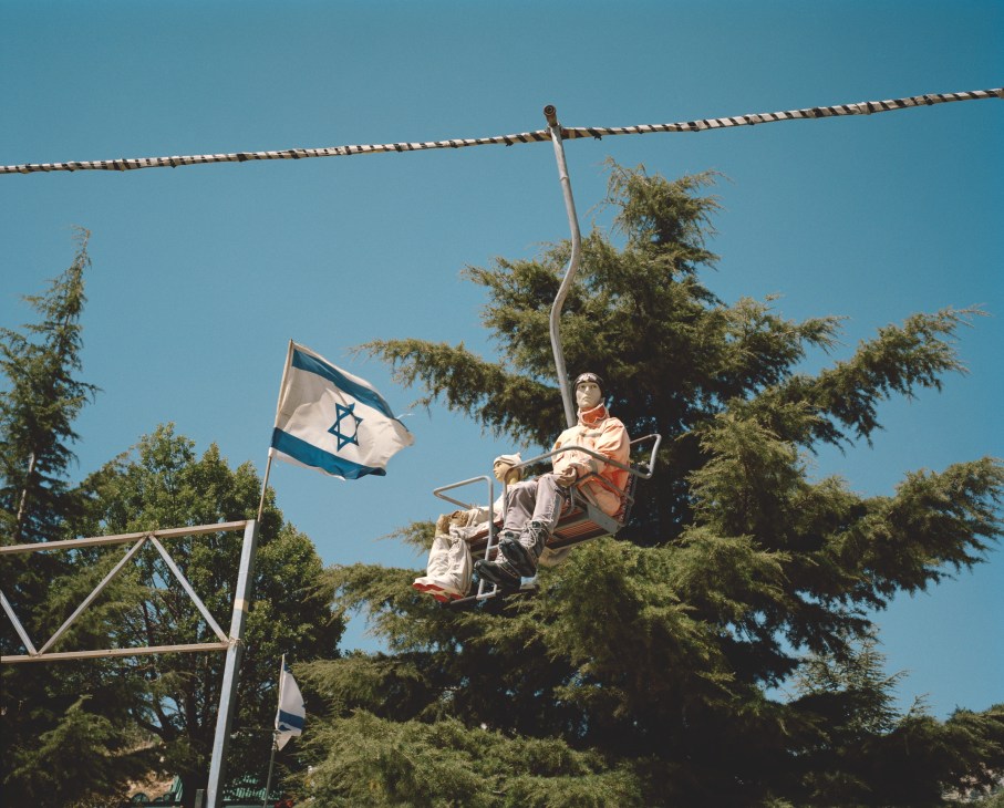 A person on a swing suspended from a tall metal structure with Israeli flags flying nearby against a clear blue sky, surrounded by coniferous trees.