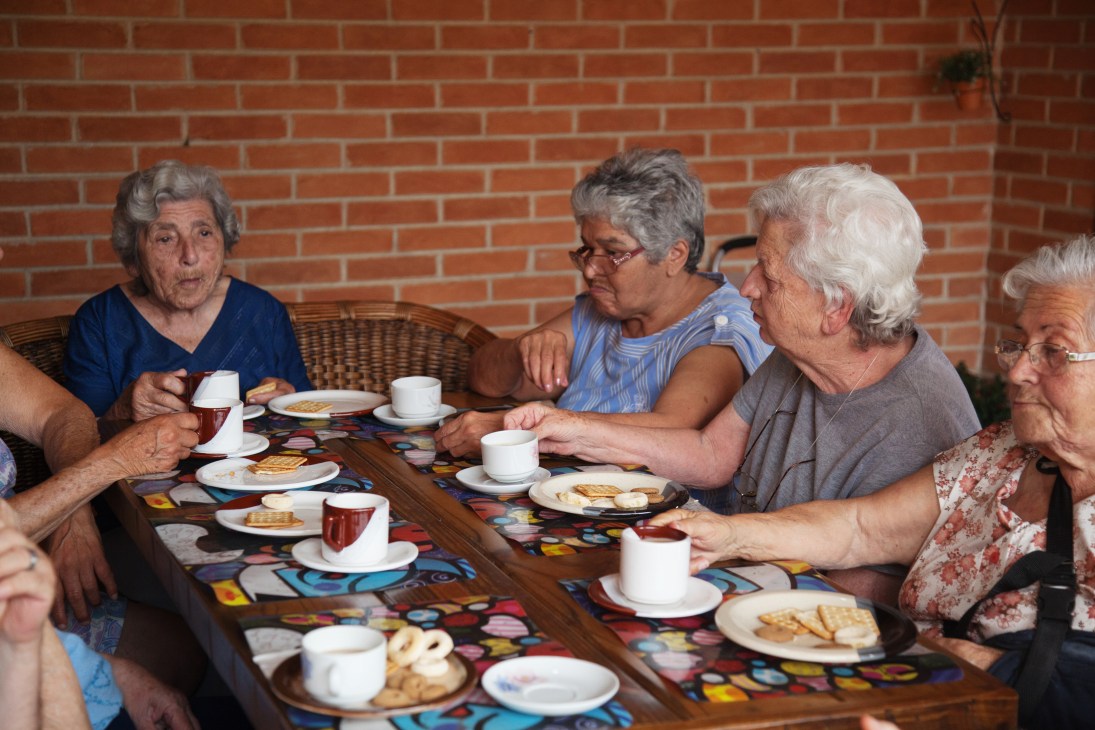 Elderly people enjoying tea and snacks together at a table with a colorful patterned tablecloth against a brick wall.