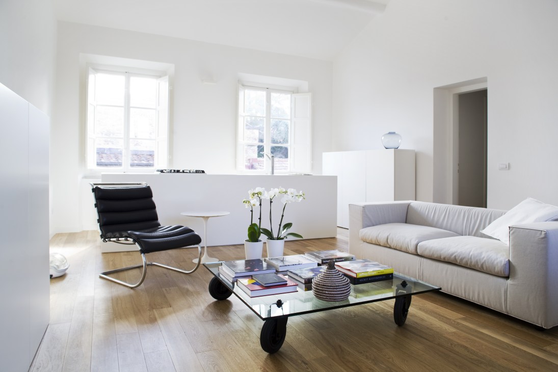 Minimalist living room with white walls, cream sofa, black chair, glass coffee table on wheels, and bright windows.