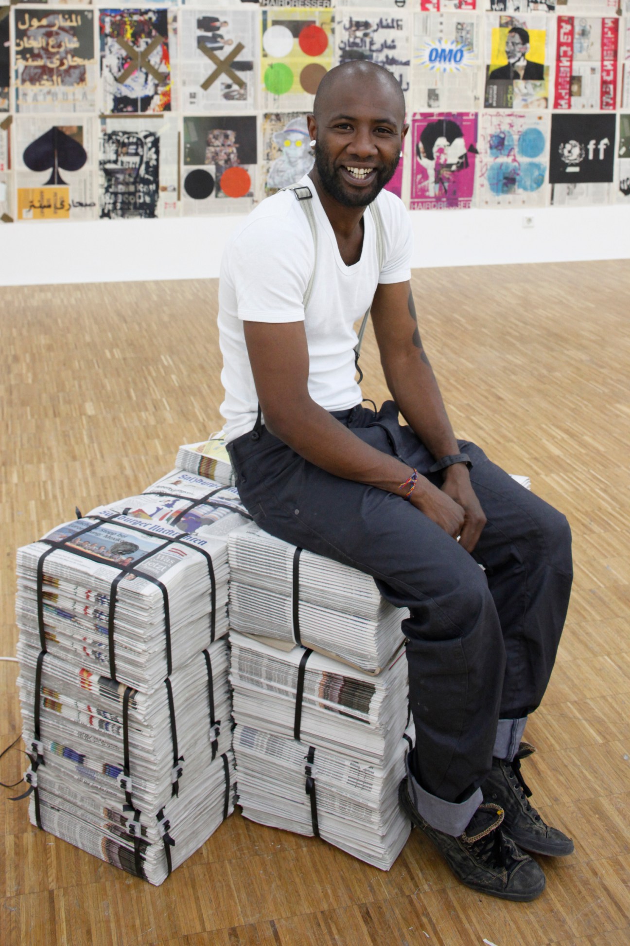 Yonamine, an Angolan artist, smiles while sitting on stacked newspapers in an art studio.