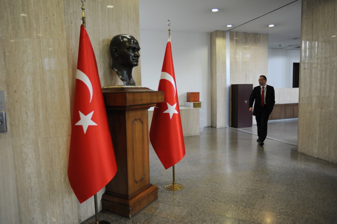 Man in dark suit walking through ministry foyer with Turkish flags and bust sculpture.
