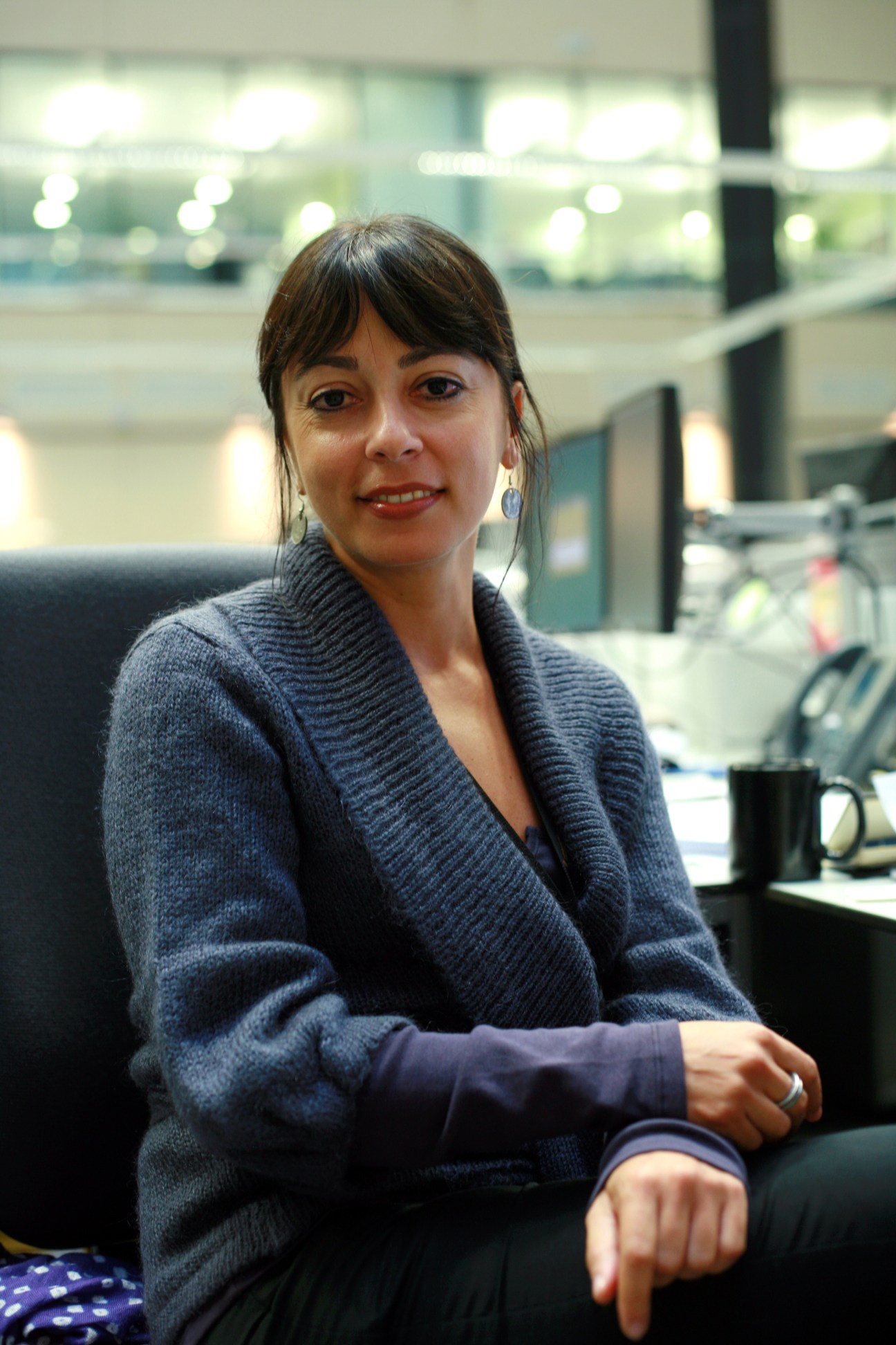 Golnaz Esfandiari, a woman with dark hair and bangs wearing a blue cardigan, smiles at the camera while seated in an RFE/RL office with glass partitions visible in the background.