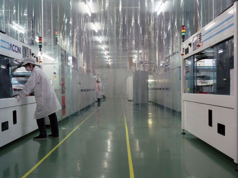 Worker in white coat operates equipment in a bright, clean laboratory with industrial machinery and status indicator lights.
